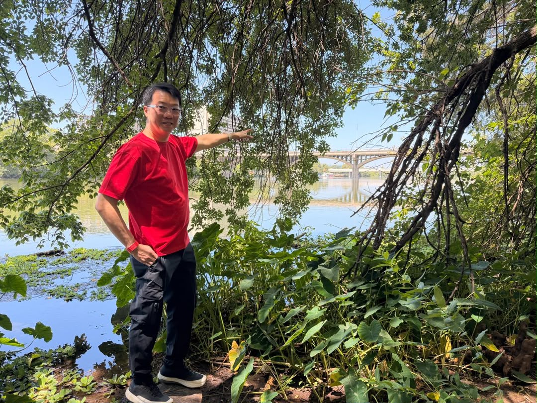 Joey Yap pointing toward a bridge crossing Austin's lake, with open water and greenery in the foreground
