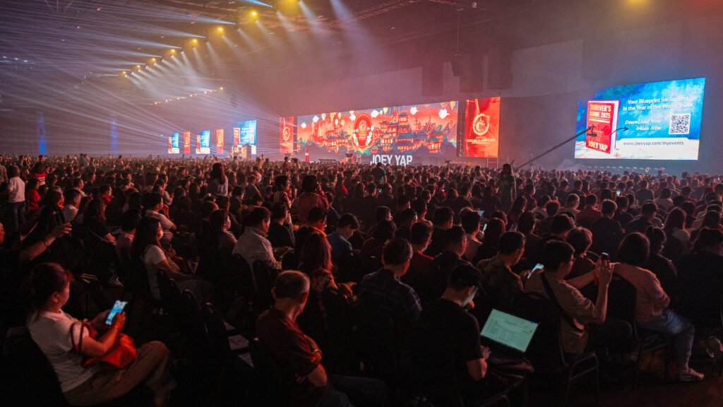 A packed audience at a Joey Yap live seminar, with the Joey Yap name displayed on the main stage screen